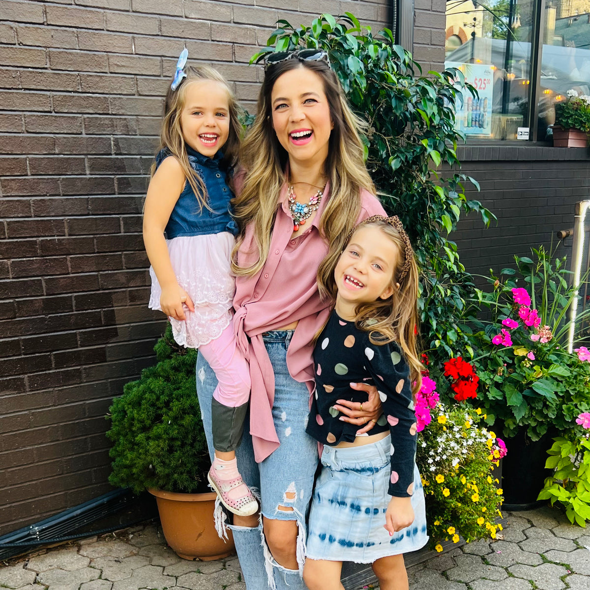 Woman with two young girls standing outdoors next to a brick wall and plants.